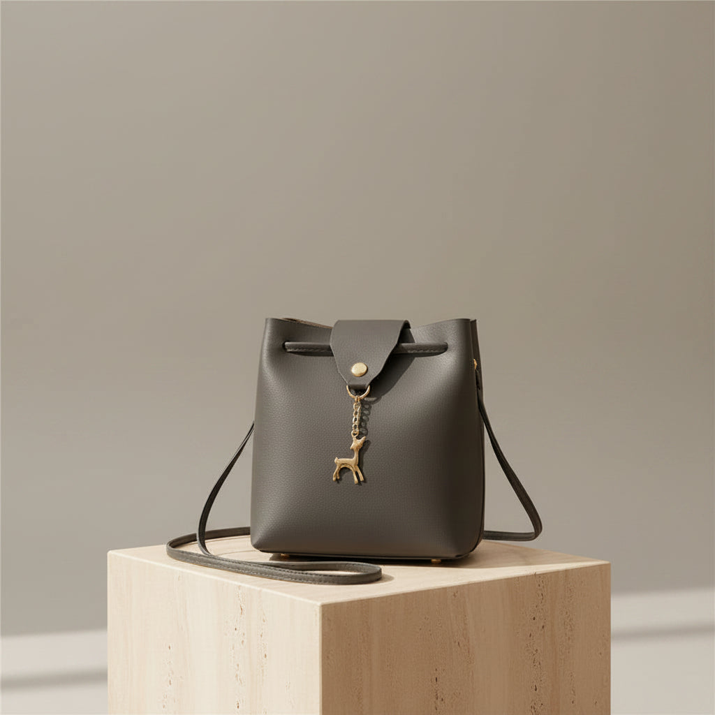 Charcoal Grey leather bucket bag with a drawstring top and a gold-tone deer charm, displayed on a light beige stone pedestal in a warm, modern interior setting.